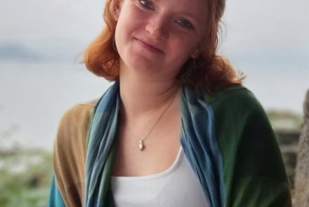 A woman stands in a white shirt and blue, green and brown coat in front of a blurred background of water.