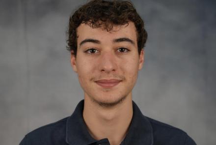 Headshot of a man with curly hair wearing a navy blue polo shirt in front of a mottled gray backdrop. 