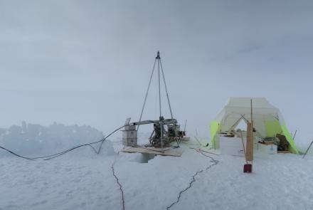 A snowy terrain. A metal drilling device rests on plywood over a hole in snow. Next to it is a white and neon yellow tent housing scientific equipment.