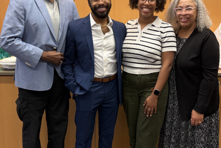Four adults stand together smiling in front of a light wood-paneled wall. From left to right: a tall man with white hair and beard wearing a light blue blazer and black pants; a man in a navy blue suit with brown shoes; a woman wearing glasses, a white striped short-sleeve sweater, and olive green pants; and a woman with curly gray hair wearing glasses, a black cardigan, and a patterned black-and-white dress. 