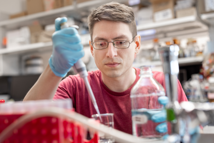 A man in a red t-shirt and glasses wears lab gloves and pipettes liquids. 