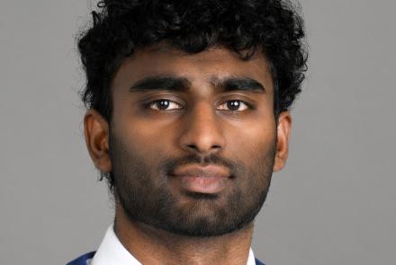 A headshot of a man in a white dress shirt, baby blue tie and royal blue sweater. He is situated in front of a gray background.