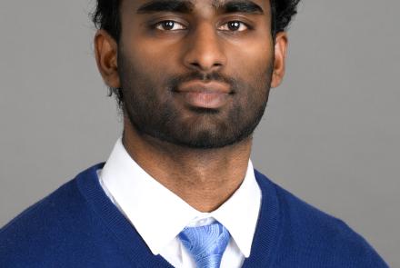Headshot of a man wearing a white dress shirt, light blue tie and royal blue sweater in front of a gray backdrop.