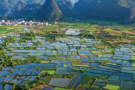 Aerial view of a patchwork of flooded rice paddies in a valley in China. In the background is a small village of white and orange buildings in front of towering karst peaks. All of this sits below a hazy sky.