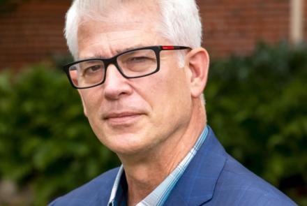 A man in black glasses and a blue suit over a blue plaid shirt stands in front of blurred greenery and bricks.