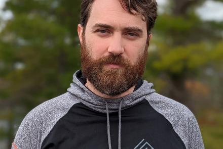 A bearded, brunette man wears a black and gray hoodie that says "SEED Electronic Arts" standing in front of a blurred background of trees.