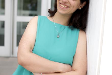 A young woman leans against a white column outdoors, smiling with her arms crossed. She wears a blue, sleeveless shirt and black pants. Glass doors are blurred in the background.
