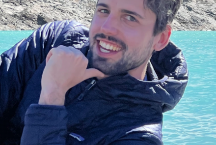 Brown haired man with beard and blue jacket smiling with his chin on his hand in front of water and rocks.