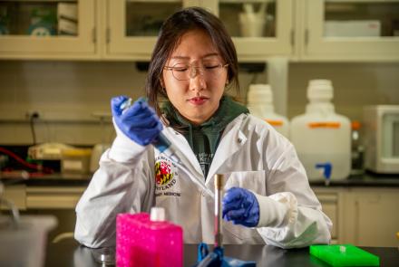 A woman in a lab coat wearing blue gloves and glasses uses a pipette to transfer liquic into a test tube.