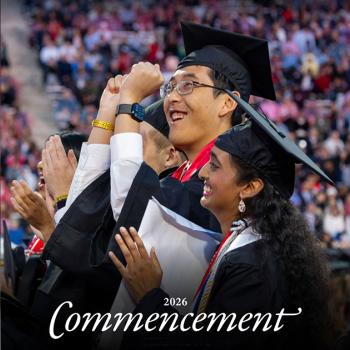 Two students in graduation regalia celebrating, with a blurred crowd in the background.