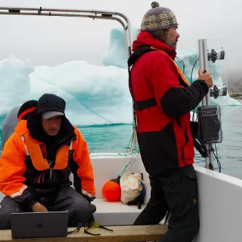 Cy Keener (seated on a boat at an iceberg in Greenland while Lars Smedsrud holds device equipped with sonar technology