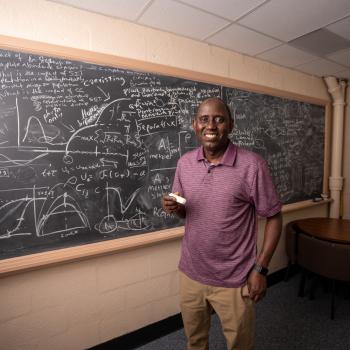 Professor Abba Gumel. wearing a red polo and khaki pants, holds a piece of chalk smiling in front of a chalkboard filled with mathematics equations and graphs. 