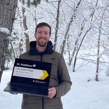 Justin Pflug wearing a coat and holding a laptop in front of a snowy forest