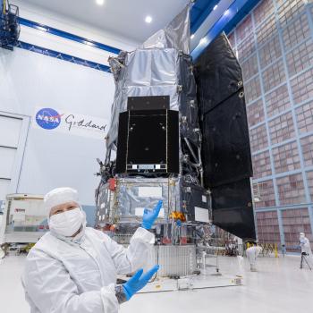 Jackie Townsend wearing all-white lab gear and blue gloves, gesturing to the Roman Space Telescope behind her