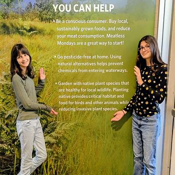 Two girls standing on either side of a wall filled with text about native plants