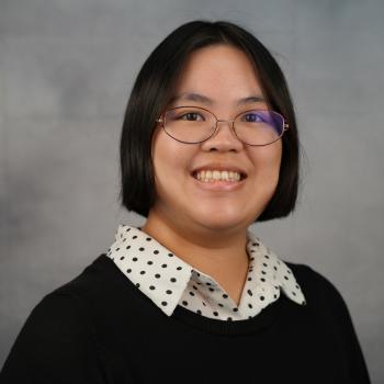 A young woman in a black-and-white polka dot button-down shirt and a black sweater smiles in front of a mottled gray backdrop.