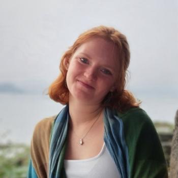 A woman stands in a white shirt and blue, green and brown coat in front of a blurred background of water.