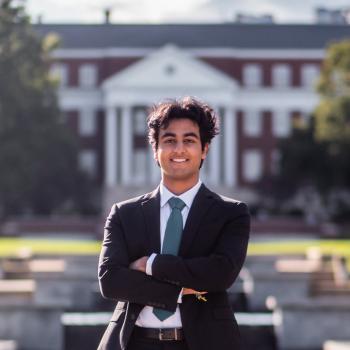 Dhruv Dewan stands in a suit in front of a red academic building.