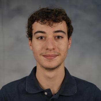 Headshot of a man with curly hair wearing a navy blue polo shirt in front of a mottled gray backdrop. 