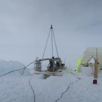 A snowy terrain. A metal drilling device rests on plywood over a hole in snow. Next to it is a white and neon yellow tent housing scientific equipment.