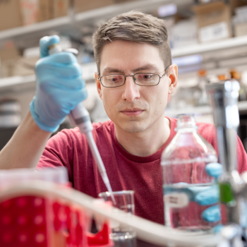 A man in a red t-shirt and glasses wears lab gloves and pipettes liquids. 