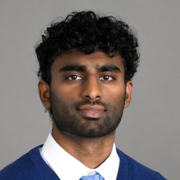 A headshot of a man in a white dress shirt, baby blue tie and royal blue sweater. He is situated in front of a gray background.