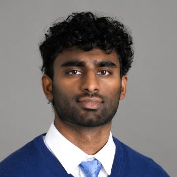 Headshot of a man wearing a white dress shirt, light blue tie and royal blue sweater in front of a gray backdrop.