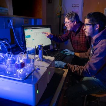 Two researchers work on a computer in a dimply lit room. One older man wears a checkered red button-down shirt, while a younger man wears a maroon hoodie. Both wear glasses. In front of them are beakers and flasks illuminated in blue light.