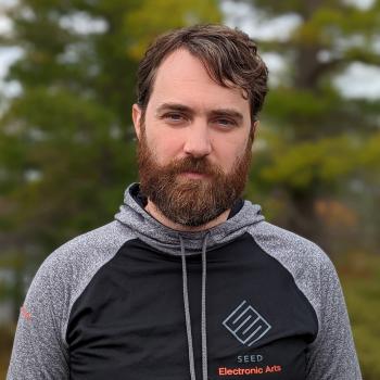 A bearded, brunette man wears a black and gray hoodie that says "SEED Electronic Arts" standing in front of a blurred background of trees.