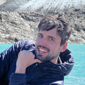 Brown haired man with beard and blue jacket smiling with his chin on his hand in front of water and rocks.