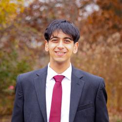 A male college-aged student wearing a black suit with a white dress shirt and red tie stands in front of a blurred background of autumnal-colored foliage.