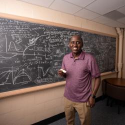 Professor Abba Gumel. wearing a red polo and khaki pants, holds a piece of chalk smiling in front of a chalkboard filled with mathematics equations and graphs. 