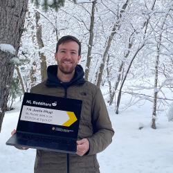 Justin Pflug wearing a coat and holding a laptop in front of a snowy forest