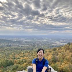 Biological sciences graduate student Stephanie Chia sits smiling at the camera on a rock overlooking a cloudy sky and a valley of trees.
