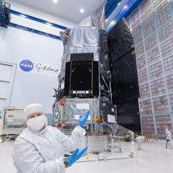 Jackie Townsend wearing all-white lab gear and blue gloves, gesturing to the Roman Space Telescope behind her