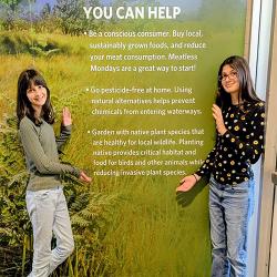 Two girls standing on either side of a wall filled with text about native plants