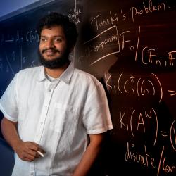 Srivatsav Kunnawalkam Elayavalli holding a piece of chalk and standing in front of a blackboard full of equations