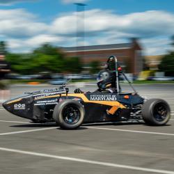 A black racecar with the text "University of Maryland" moves along a blurry background of a blue, cloudy sky and green trees.