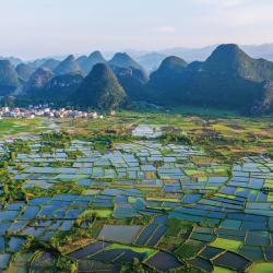 Aerial view of a patchwork of flooded rice paddies in a valley in China. In the background is a small village of white and orange buildings in front of towering karst peaks. All of this sits below a hazy sky.