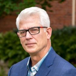 A man in black glasses and a blue suit over a blue plaid shirt stands in front of blurred greenery and bricks.