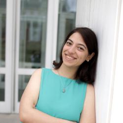 A young woman leans against a white column outdoors, smiling with her arms crossed. She wears a blue, sleeveless shirt and black pants. Glass doors are blurred in the background.
