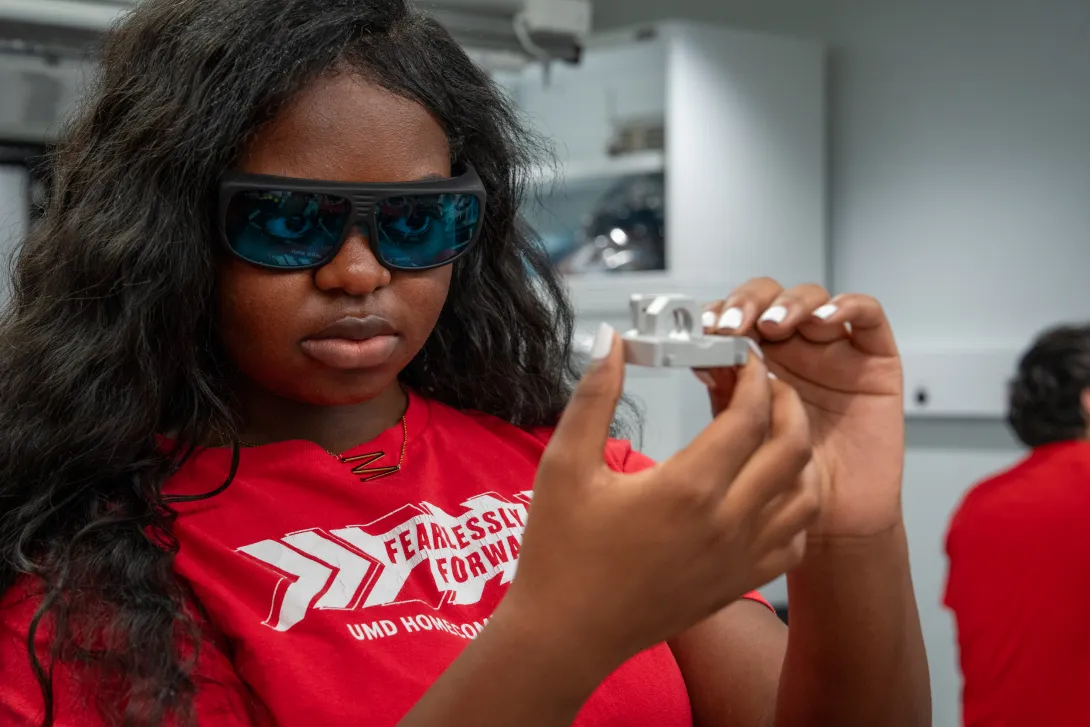 A student wearing lab safety glasses and a red shirt holding something up