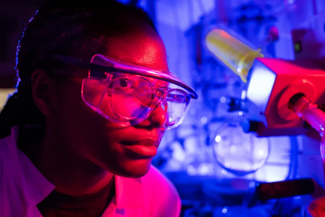 A close-up portrait of a researcher wearing safety goggles, lit in dramatic red and blue tones, with laboratory glassware and equipment blurred in the background.