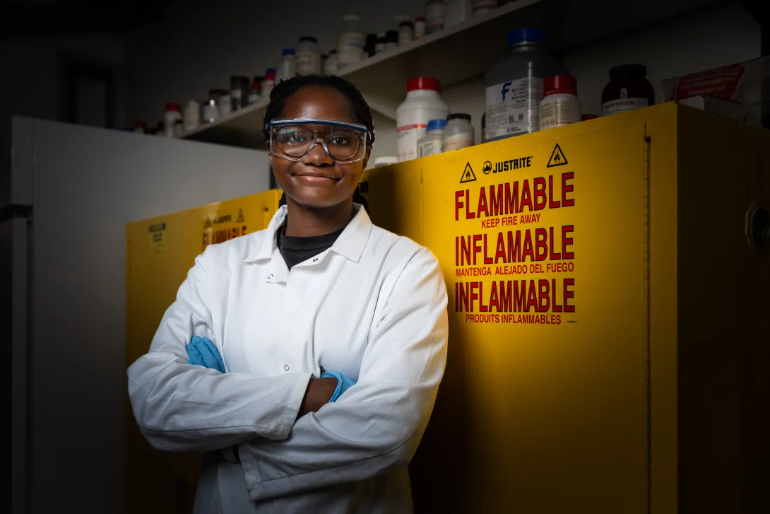 A researcher in a white lab coat, safety goggles, and gloves stands with arms crossed in front of yellow flammable-chemical storage cabinets lined with reagent bottles.