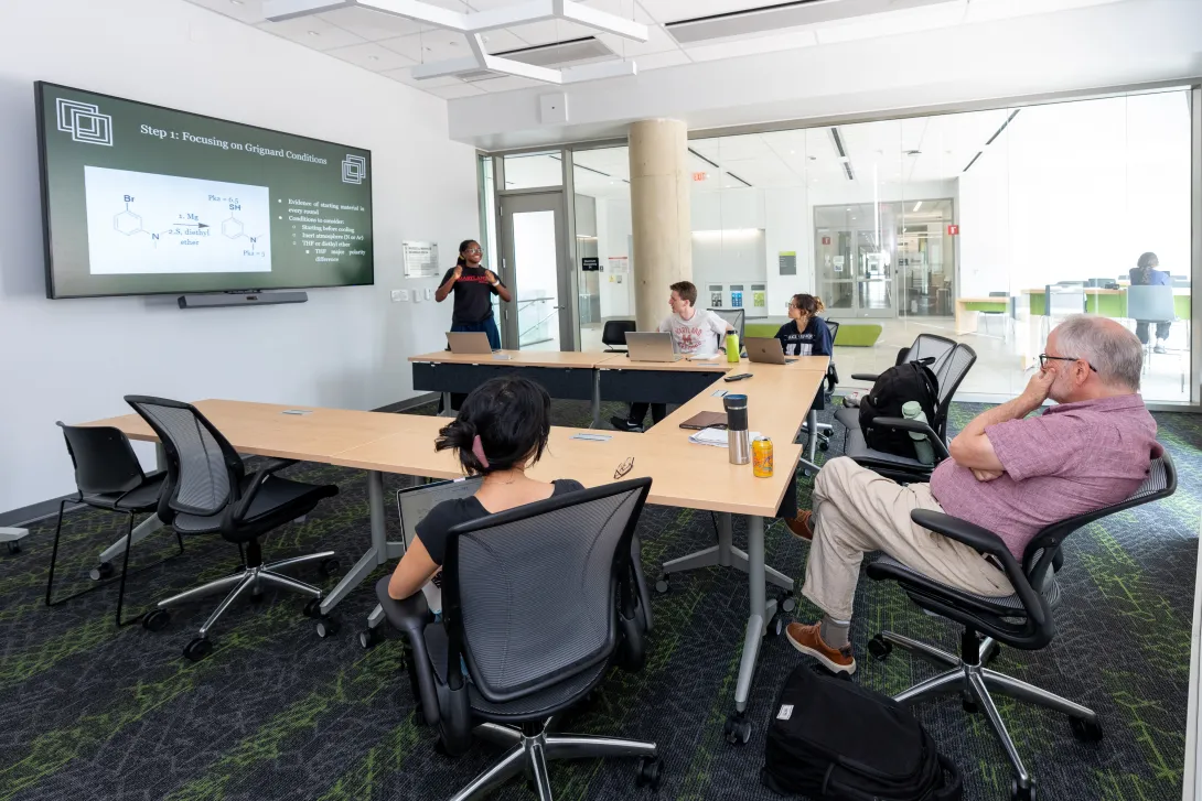 A researcher presents a slide on Grignard reaction conditions to colleagues seated around tables in a seminar room, with organic chemistry structures displayed on a large screen.
